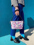 Small vegan leather crossbody purse with a real pebble grain texture and adjustable black strap. The flat printed artwork features a scattered all over pattern of pink skulls, purple and pastel blue winged bats, pink fanged mouths, and loose eyeballs set against a cream colored background. The bag is shown with black and silver hardware including a black top zipper, black zipper pull, black strap tabs, and silver metal rings and clips. The compact rectangular bag measures 8.5 by 7 by 2 inches.