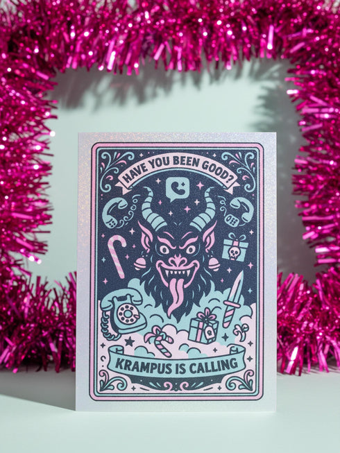 A vertical 5x7 holiday card featuring a retro tattoo style illustration of Krampus in pastel pink, teal, and black ink on shimmery silver pearlescent paper. The central horned demon has a long tongue and is surrounded by vintage rotary phones, daggers, candy canes, and skull gifts. Top text asks Have You Been Good while the bottom banner reads Krampus Is Calling. The interior text reads Let's be honest... Being good is overrated. Merry Christmas.