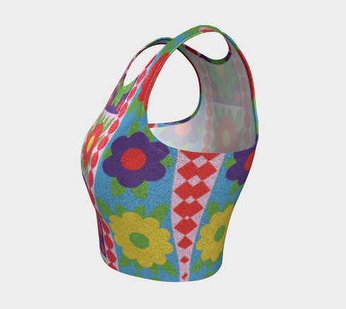 A high-resolution product photo of a body-hugging, 2D printed crop top with a 1970s-inspired floral patchwork pattern. The design consists of vibrant squares in cobalt blue, pale lavender, and deep violet, separated by pink borders with red dots. Central motifs include yellow daisies with orange centers and green-magenta floral mandalas. The top is made of a smooth, high-stretch performance knit fabric with curved back armholes and a cropped waistline.