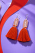 A pair of red tassel earrings with a tassel at the end, displayed on a purple background with a red and purple curved line.