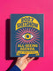 A hardcover spiral planner for 2027 with a dark blue cover and retro 70s style graphics. The title reads "2027 SHITSHOW All-Seeing Agenda" in bold yellow and pink text. Below the title is a large, stylized all-seeing eye with pink rays emanating from it. At the bottom, a pink ribbon contains the text "BLINK TWICE FOR MERCY." The back cover is also dark blue with a yellow sun graphic and the text "Plans bloom. Deadlines hum. I write, I wander, then get it done. Keep receipts. Keep grace.