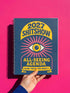 A hardcover spiral planner for 2027 with a dark blue cover and retro 70s style graphics. The title reads "2027 SHITSHOW All-Seeing Agenda" in bold yellow and pink text. Below the title is a large, stylized all-seeing eye with pink rays emanating from it. At the bottom, a pink ribbon contains the text "BLINK TWICE FOR MERCY." The back cover is also dark blue with a yellow sun graphic and the text "Plans bloom. Deadlines hum. I write, I wander, then get it done. Keep receipts. Keep grace.