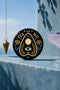 A close-up photograph of a glossy ceramic coaster featuring a Planchette occult design on a deep black background. The symmetrical graphic displays a central blue All-Seeing Eye with gold sunburst lines, topped by a white circle, and flanked by gold flowers with blue stems. At the top, the text "YES" is printed in gold on the left and "NO" on the right. Gold stars and crescent moons are scattered around the perimeter. The flat, round coaster has a highly visible high-gloss glaze.