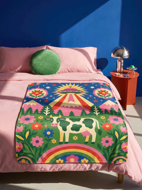 A rectangular velveteen minky blanket with a bright, maximalist design. The top half features a vibrant blue sky with a pink, green, and yellow UFO beaming rays down over hot pink snow-capped mountains. The bottom half shows a grassy green field with a white cow covered in dark spots, surrounded by large, colorful red, pink, and blue retro flowers, pine trees, and a rainbow arch at the very bottom. The reverse side is a fluffy off-white sherpa.