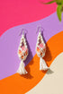 A pair of white earrings with a floral design, hanging from hooks against a colorful background.