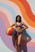 Two piece high waisted bikini in a bold 70s psychedelic geometric print. The sporty scoop top features bright yellow and red sunbursts over a black, purple, cream, and red colorblock background. The high cut bottoms carry matching geometric diamonds with hot pink and teal abstract palm leaves. Smooth form fitting recycled polyester fabric with UPF 50 sun protection and removable padding. Vibrant maximalist retro swimwear in saturated primary and secondary shades, made for pool parties, festivals, and loud s