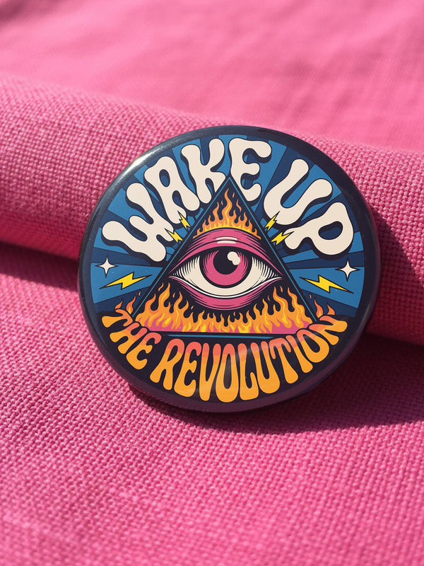 A round pinback button featuring a retro psychedelic design. The central focus is a stylized Eye of Providence in a pink triangle, surrounded by vibrant orange and yellow flames. Behind the triangle are teal and petrol blue sunburst rays. The text "WAKE UP" curves across the top in cream wavy letters, and "THE REVOLUTION" curves along the bottom in orange. The button has a glossy finish and is available in 1.75 inch or 2.25 inch sizes.