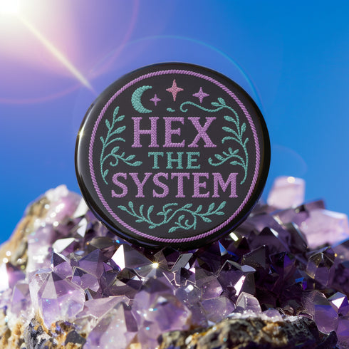Glossy black pinback button with pastel embroidery-style design that says “Hex the System,” surrounded by vines, stars, and a crescent moon. Displayed against witchy pink and blue backdrops, street pole flyers, and protest props.