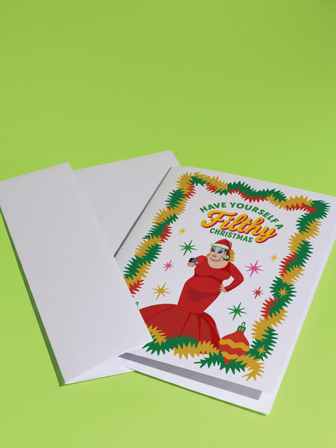 A 5x7 pearlescent holiday card by HoneyDazed. The card illustration, inspired by John Waters, features the drag queen Divine in a red dress with a Santa hat. She points a gun, withtext reads "Have Yourself a Filthy Christmas" in campy font. The card is bordered by garland. Iinside of the card reads: "Season's Sleazings" in red and green text. This kitschy greeting card is a perfect piece of cult film memorabilia and a funny adult humor gift for fans of queer art.