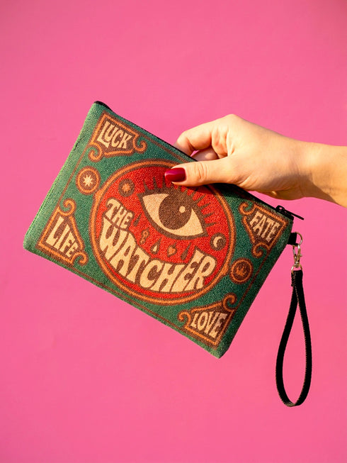 A rectangular 9x6 inch makeup bag and zipper pouch featuring a textured linen-like polyester fabric. The bag showcases a bold, maximalist print with vivid colors typical of retro 70s and pastel goth aesthetics, centering on a large mystic eye design labeled "The Watcher." It features a sturdy black zipper across the top and includes a removable black nylon wristlet strap.Perfect for use as a pencil case, cosmetic organizer, or festival wristlet.