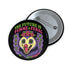 A round, black pinback button with a high-gloss finish features the text "THE FUTURE IS FEMME & FERAL" in a retro font of lime green and pastel pink. In the center, a screaming possum with wild eyes and a pink-lined mouth is illustrated in a pastel goth style with purple, yellow, and pink fur. The possum wears a pink flower crown. The illustration is framed by a circle of small skulls and pink Venus (female) symbols on a purple background. This is a piece of feminist possumcore flair.