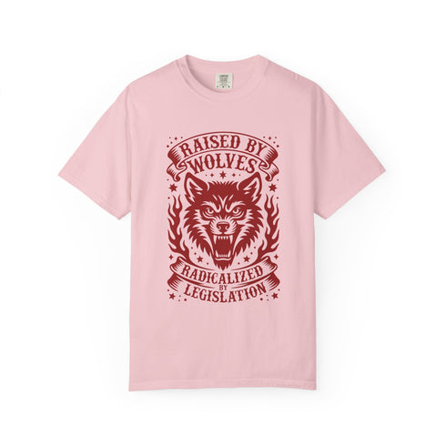 Vintage style graphic t-shirt with a large snarling wolf face printed in dark charcoal ink. A curved banner above reads RAISED BY WOLVES and a banner below reads RADICALIZED BY LEGISLATION, framed by stylized flames and scattered stars. Classic crewneck short sleeve cotton tee with a relaxed fit and seamless sides, available in soft pink, pale green, and ivory. Feminist protest apparel and activist statement tee for reproductive rights, marches, and everyday quiet rage.