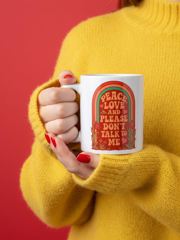 A glossy white porcelain ceramic mug featuring a vibrant maximalist design centered on the exterior. The artwork displays a retro 1970s-inspired rainbow arch composed of concentric bands in burnt orange, mustard yellow, and teal green. Inside the arch, stylized vintage bubble-serif typography in reddish-orange reads PEACE LOVE AND PLEASE DON'T TALK TO ME. The design evokes a groovy, hippie aesthetic mixed with sassy introvert humor.