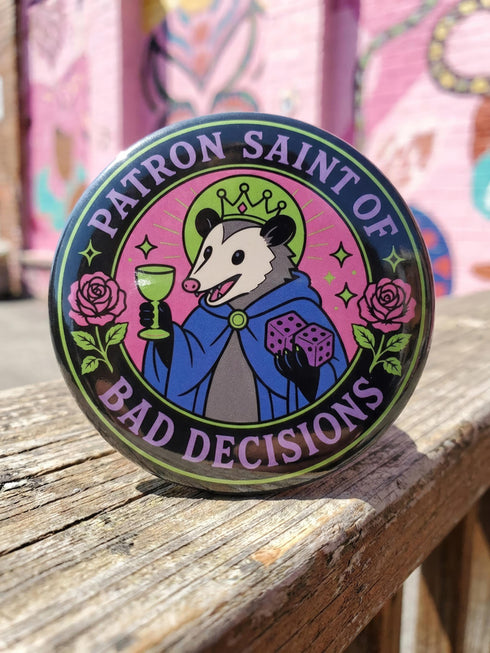 A round glossy metal pinback button featuring a flat 2D print illustration of an opossum wearing a blue cloak and a green crown. The opossum holds a neon green goblet in one paw and two purple dice in the other. A bright pink circle sits behind the opossum, surrounded by a black outer ring. Purple text reads PATRON SAINT OF BAD DECISIONS curved around the edge. Two pink roses with green leaves are on the sides. The design uses bold black outlines and bright neon colors.