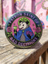 A round glossy metal pinback button featuring a flat 2D print illustration of an opossum wearing a blue cloak and a green crown. The opossum holds a neon green goblet in one paw and two purple dice in the other. A bright pink circle sits behind the opossum, surrounded by a black outer ring. Purple text reads PATRON SAINT OF BAD DECISIONS curved around the edge. Two pink roses with green leaves are on the sides. The design uses bold black outlines and bright neon colors.