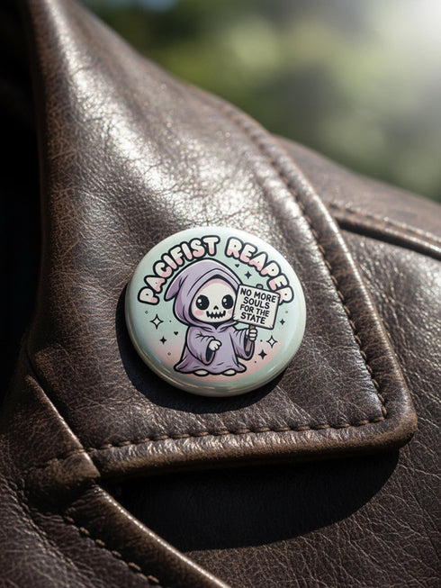 Round pinback button featuring a pastel mint green background with a cartoon grim reaper character in a lavender hood holding a protest sign. The design includes bold black outlined lettering reading “Pacifist Reaper” above the figure and small sparkle accents surrounding it. The finish is glossy and smooth, with a printed illustration style on a metal pin badge surface.