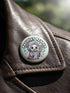 Round pinback button featuring a pastel mint green background with a cartoon grim reaper character in a lavender hood holding a protest sign. The design includes bold black outlined lettering reading “Pacifist Reaper” above the figure and small sparkle accents surrounding it. The finish is glossy and smooth, with a printed illustration style on a metal pin badge surface.