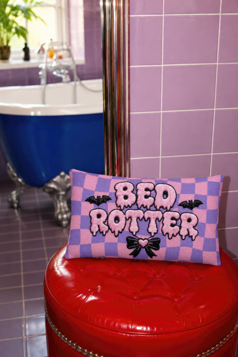 Pastel goth lumbar pillow, 20 by 12 inches, with a pink and lavender purple wavy checkerboard background. The words Bed Rotter appear in dripping pink bubble letters with a faux embroidery thread texture and black outlines. Two black bats with pink stitch details flank the text, and a black bow with a pink heart center sits below. Double sided flat print that mimics raised embroidery on linen feel polyester with a hidden zipper, insert included.