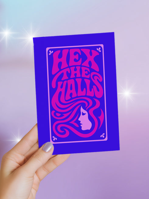 A vertical 5x7 greeting card made from premium 120lb silk cover stock with a slight sheen. The card has a deep purple background with a thin pink border. In the center, the words 'Hex the Halls' are written in a bubbly, psychedelic 70s-style font in shades of hot pink and light pink. Below the text is a stylized pink silhouette of a woman's face in profile with long, flowing, wavy hair. The design is reminiscent of retro witchcraft and groovy aesthetics, perfect for an alternative holiday.
