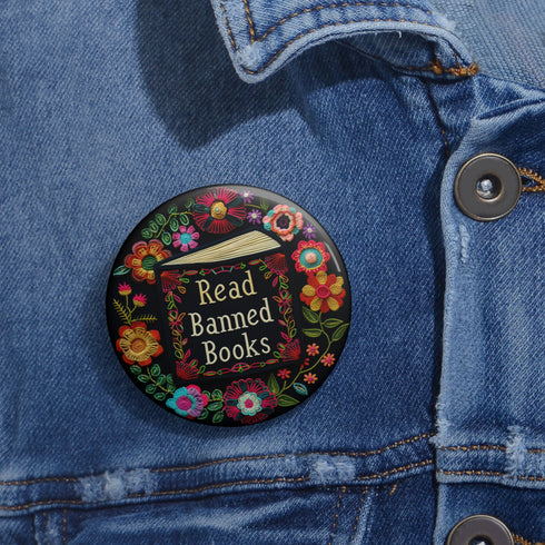 Round black pinback button with "Read Banned Books" in gold lettering on an open book image, surrounded by vibrant embroidery-style flowers in pink, orange, blue and green. Folk art-inspired literary protest pin with glossy finish and safety pin backing, available in 2" and 3" sizes.