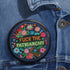 Circular black pinback button with "FUCK THE PATRIARCHY" in orange and mint green text, surrounded by embroidery-style flowers in pink, blue, and red with green foliage and blue border. Folk art feminist protest pin with glossy finish and safety pin backing in 2" and 3" sizes.