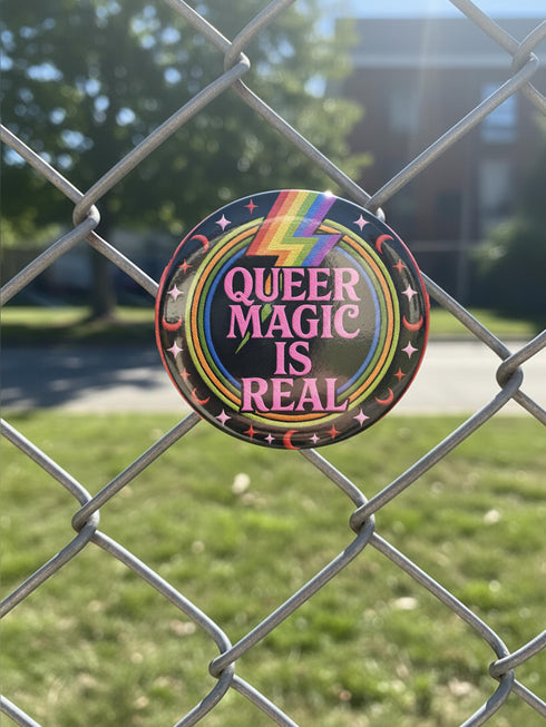 A round metal pinback button badge featuring a black background with the words QUEER MAGIC IS REAL in capital pink retro serif typography. A bright rainbow colored lightning bolt strikes from the top center, surrounded by concentric rainbow rings in red, orange, yellow, green, blue, and purple. The design is accented with small pink four-point stars and red crescent moons. The artwork features a realistic 2-D faux embroidery texture. The back has a silver metal safety pin clasp fastening.