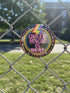 A round metal pinback button badge featuring a black background with the words QUEER MAGIC IS REAL in capital pink retro serif typography. A bright rainbow colored lightning bolt strikes from the top center, surrounded by concentric rainbow rings in red, orange, yellow, green, blue, and purple. The design is accented with small pink four-point stars and red crescent moons. The artwork features a realistic 2-D faux embroidery texture. The back has a silver metal safety pin clasp fastening.