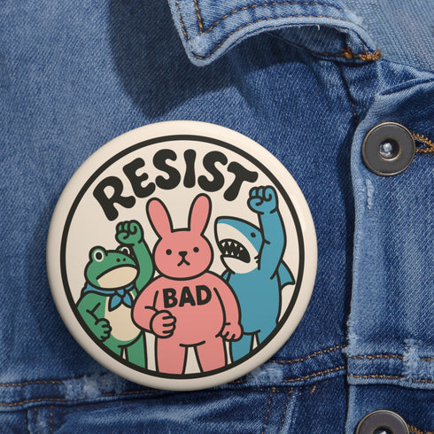 A round white pinback button with the word 'RESIST' in a bold, black, arched font. Below the text stand three cute cartoon characters raising their fists in protest: a green frog with a blue bandana, a pink bunny with '/BAD' on its chest, and a blue shark with sharp teeth. The activist pin has a glossy, protective mylar finish and a durable metal shell, designed as political statement flair for jackets, bags, and backpacks. This punk-inspired, kawaii- badge is a symbol of joyful rebellion.