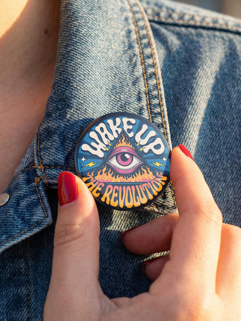A round pinback button featuring a retro psychedelic design. The central focus is a stylized Eye of Providence in a pink triangle, surrounded by vibrant orange and yellow flames. Behind the triangle are teal and petrol blue sunburst rays. The text "WAKE UP" curves across the top in cream wavy letters, and "THE REVOLUTION" curves along the bottom in orange. The button has a glossy finish and is available in 1.75 inch or 2.25 inch sizes.