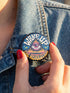 A round pinback button featuring a retro psychedelic design. The central focus is a stylized Eye of Providence in a pink triangle, surrounded by vibrant orange and yellow flames. Behind the triangle are teal and petrol blue sunburst rays. The text "WAKE UP" curves across the top in cream wavy letters, and "THE REVOLUTION" curves along the bottom in orange. The button has a glossy finish and is available in 1.75 inch or 2.25 inch sizes.