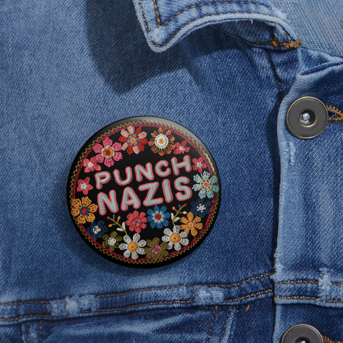 Round pinback metal button featuring an embroidered-style design on a black background. The center showcases bold, stitched-textured lettering in red, blue, and white that reads "PUNCH NAZIS." Surrounding the text is an intricate, folk-inspired floral pattern with multicolored flowers in shades of pink, orange, blue, and white, arranged symmetrically. A decorative zigzag border frames the edge of the button. The backside is silver metal with a secure pin closure.