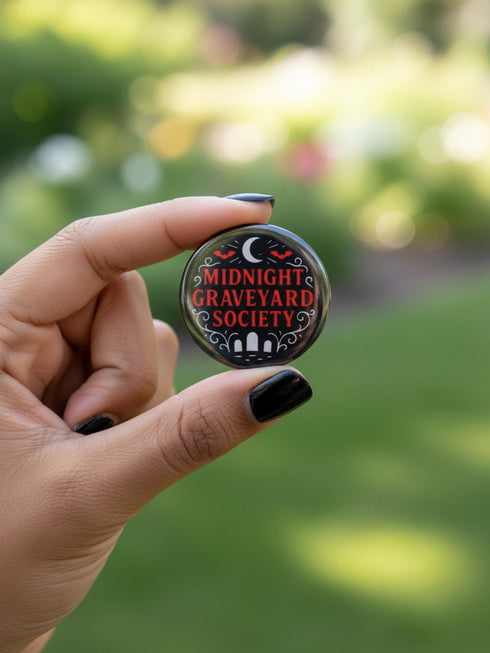 A collection of four glossy black circular pinback buttons featuring striking red and white gothic illustrations. One button shows "Midnight Graveyard Society" text with bats and tombstones. Another reads "Cemetery Picnic Society" with a coffin and umbrella icon. A third features a detailed white skull and red roses with "Memento Mori" text below. The final pin displays "Nocturnal Flower Society" surrounded by floral vines.