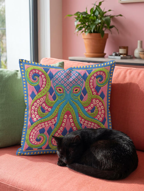 A square decorative throw pillow cover featuring a highly symmetrical bright octopus design. The octopus has a turquoise blue body with a diamond patterned head and eight curled tentacles detailed in bright pink and lime green. The background is a repeating diamond argyle pattern in royal blue and light pink. The artwork is a flat two dimensional print that mimics the textured look of intricate crewelwork embroidery. The pillow features a hidden zipper on the bottom edge. No insert included.