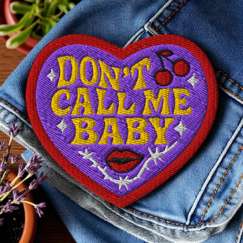 A heart-shaped embroidered patch with a thick red border and a vibrant purple background. In the center, the phrase "DON’T CALL ME BABY" is stitched in a bold, golden-yellow block font. The patch is decorated with small white sparkles, a pair of red cherries with a black musical note, a pair of red lips, and a grey thorny vine design below the text. The overall design has a rebellious, retro, punk aesthetic with a handmade, textured look.