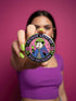 A round glossy metal pinback button featuring a flat 2D print illustration of an opossum wearing a blue cloak and a green crown. The opossum holds a neon green goblet in one paw and two purple dice in the other. A bright pink circle sits behind the opossum, surrounded by a black outer ring. Purple text reads PATRON SAINT OF BAD DECISIONS curved around the edge. Two pink roses with green leaves are on the sides. The design uses bold black outlines and bright neon colors.