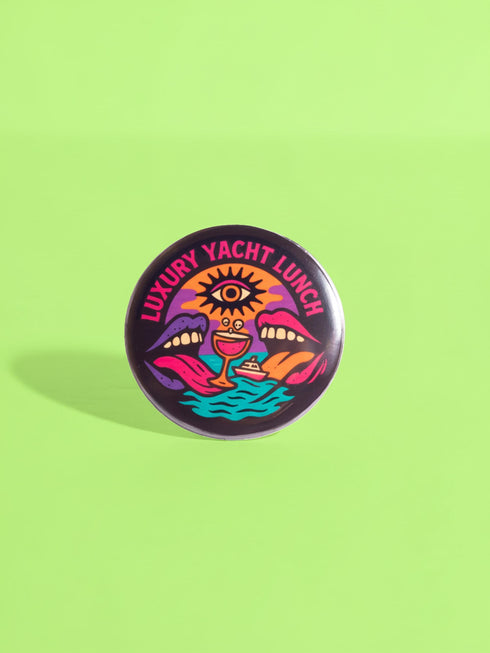 A round 1.75 inch glossy pinback button featuring satirical psychedelic artwork on a black background. The text LUXURY YACHT LUNCH arches over the top in bold neon pink letters. The central design depicts a surreal scene with a stylized third-eye sun, large open mouths with tongues sticking out, a smiling cocktail glass character, and a small white yacht floating on wavy teal water. The color palette includes retro 70s neon pink, orange, purple, and teal. The button has a safety pin backing.