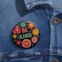 Bold "BE KIND" pinback button featuring vibrant psychedelic flowers in hot pink, teal, yellow, and orange on black background. Retro 70s-inspired typography in coral and mint green surrounded by colorful blooms. Statement activist accessory perfect for punk fashion, protest wear, and battle vests. Glossy UV-resistant finish with safety pin backing available in 2" and 3" sizes.