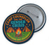 Round glossy pinback button measuring 2.25 inches featuring a playful camp-themed design with the phrase “Camp Counselor for the Gender Crisis.” The artwork includes two orange tents, a rainbow campfire, stars, and green grass. Bottom text reads “Cabin Queer, Unit Psychedelic.” Made from durable tinplate with a safety pin back and coated in a UV and scratch-resistant mylar finish. Bold and colorful, perfect for jackets, bags, or pride outfits.