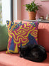 A square decorative throw pillow cover featuring a trippy retro Bigfoot scene. A large navy blue silhouette of Sasquatch walks through a forest of navy and yellow pine trees. The background is a psychedelic swirling sky in vibrant pink and yellow, featuring a large stylized eye and a cluster of small yellow stars. The flat two dimensional print visually mimics the textured look of heavy needlepoint embroidery. The pillow features a sleek hidden zipper on the bottom. No insert is included.