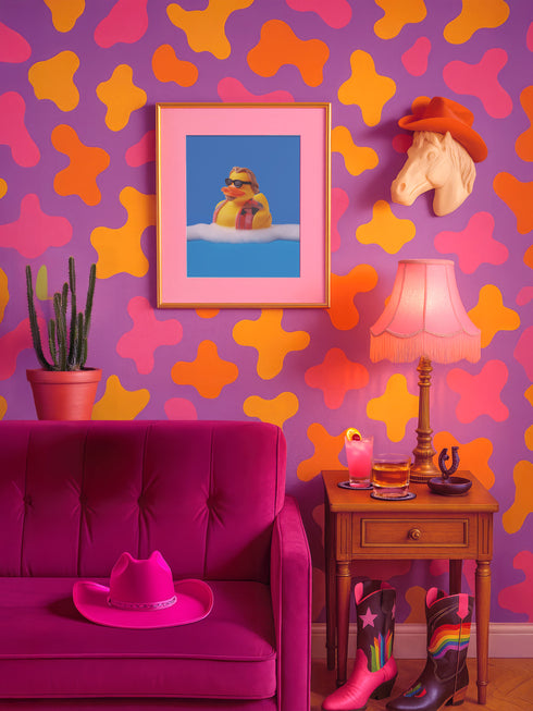 A quirky art print of a rubber duck styled after The Dude from The Big Lebowski, complete with a goatee, sunglasses, and plaid bathrobe. The duck lounges in bubbles, framed in a glossy red frame, with a manicured hand adjusting it against a monochromatic red backdrop.