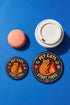 Glossy mylar pinback button featuring a solid black background. The central illustration shows a speckled orange cat sitting beside a stylized yellow and red flame. Bold retro text reads PET CATS, curved at the top, and START FIRES curved at the bottom. The design is framed by a colorful folk art border containing purple and red flowers, green leaves, and two floating eyes with light blue irises. The physical button is perfectly round with a highly reflective surface and a sturdy pin back.
