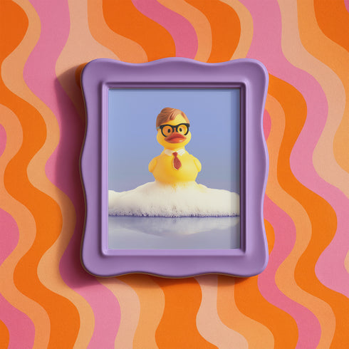 A humorous fine art print of a rubber duck modeled after Dwight Schrute, complete with glasses, mustard tie, and serious expression. Floating in soap suds, the print is framed above a retro bed with bold orange flower wallpaper and bright accent pillows.