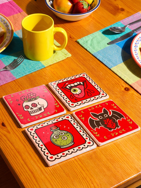 Set of four square glazed ceramic coasters featuring a vibrant pastel goth and creepy cute design. These functional art pieces show a spooky yet playful aesthetic perfect for alternative home decor. The coasters have a glossy wipe-clean finish and a non-slip cork backing to protect furniture surfaces from hot and cold beverages. Ideal for maximalist coffee tables, witchy desks, or as a unique housewarming gift for horror fans who love retro 70s and dopamine decor styles.