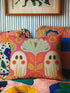 a colorful bed with a pink pillow featuring a floral design and two ghostly figures. The pillow is placed on a bed with a quilt and other pillows, creating a cozy and whimsical atmosphere.