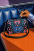 Small vegan leather crossbody purse with a real pebble grain texture and adjustable black strap. The flat printed artwork features a central blue sunburst evil eye motif below a coral pink sacred heart eye, surrounded by pink crescent moons, multicolored stars, flowing teal botanical vines, and wavy lavender and black checkerboard distortion banners. Set against a dark background with silver metal hardware including a top zipper, zipper pull, and strap clips. Measures 8.5 by 7 by 2 inches.