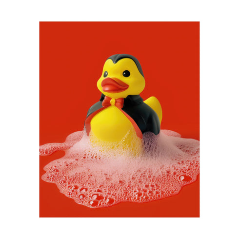 A spooky-yet-cute art print of a vampire-themed rubber duck dressed in a red-lined black cape and bowtie. Surrounded by white bath bubbles, the duck is framed in a red wavy-edged frame and held up against a burnt orange wall by a hand in a dramatic red sleeve.