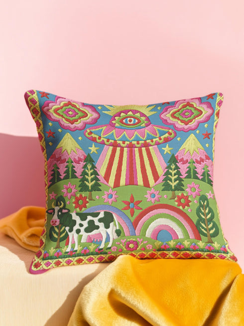 Description of a square throw pillow with a vibrant maximalist alien design. A central UFO with a large all-seeing eye hovers above a landscape with two rainbows, pink mountains, and green pine trees. A black and white spotted cow stands in the foreground. The design is a high-contrast palette of sky blue, magenta, hot pink, and meadow green. The pillow has a faux-embroidery 2D print finish on linen-feel fabric, framed by a yellow and pink scalloped floral border.