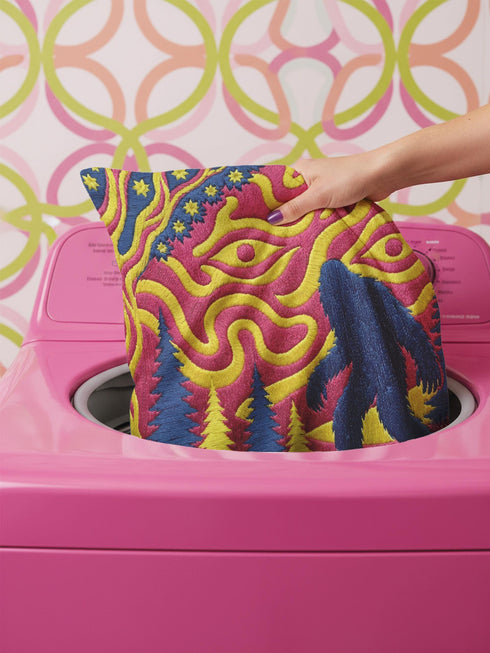A square decorative throw pillow cover featuring a trippy retro Bigfoot scene. A large navy blue silhouette of Sasquatch walks through a forest of navy and yellow pine trees. The background is a psychedelic swirling sky in vibrant pink and yellow, featuring a large stylized eye and a cluster of small yellow stars. The flat two dimensional print visually mimics the textured look of heavy needlepoint embroidery. The pillow features a sleek hidden zipper on the bottom. No insert is included.
