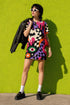 A short-sleeve, above-the-knee t-shirt dress with a relaxed, straight fit and a crew neckline. The dress features a vibrant, chaotic, all-over graphic print. The design includes sections of distorted, wavy black and cream checkerboard, mixed with bold, abstract shapes in bright purple, hot pink, and vivid orange. Scattered throughout the pattern are motifs of hot pink dripping slime, a black bat silhouette, a stylized green eye with a pink iris, and retro pink and purple daisy flowers.