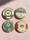 Four 2.25-inch circular metal pinback buttons on a pink textured background. The designs feature vintage tattoo-style art in rust, ochre, gold, and teal with heavy black linework. Designs include a snake Ouroboros "BREAK THE CYCLE," an umbrella eye "PROTECT THE VULNERABLE," a staircase "THE FUTURE IS OURS," and a radial eye "TRUST YOUR GAZE." The pins have a high-gloss finish. Perfect accessory for backpacks, jackets, and lanyards.